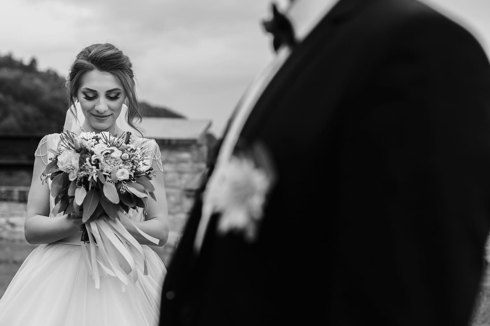 Black and white wedding photo of a bride smiling while holding her bouquet, with the groom blurred in the foreground