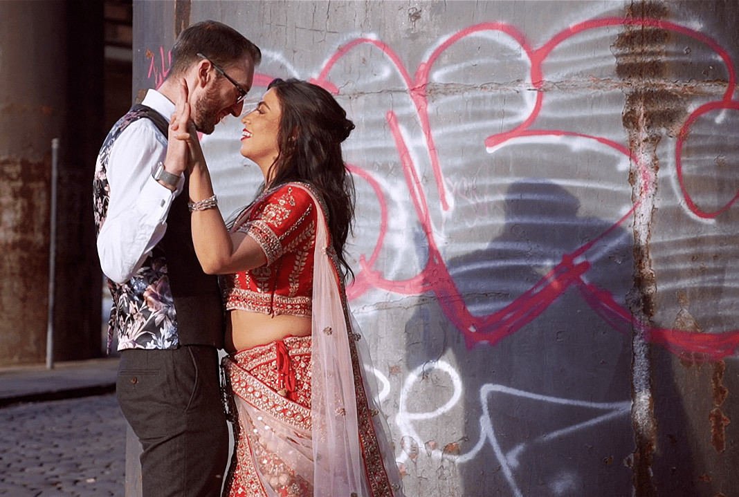 Couple Dancing Against Graffiti Wall – Manchester City Centre Castlefield Wedding Bride in red lehenga and groom in waistcoat share a joyful moment dancing against a graffiti-covered wall in a vibrant Manchester city centre wedding castlefield